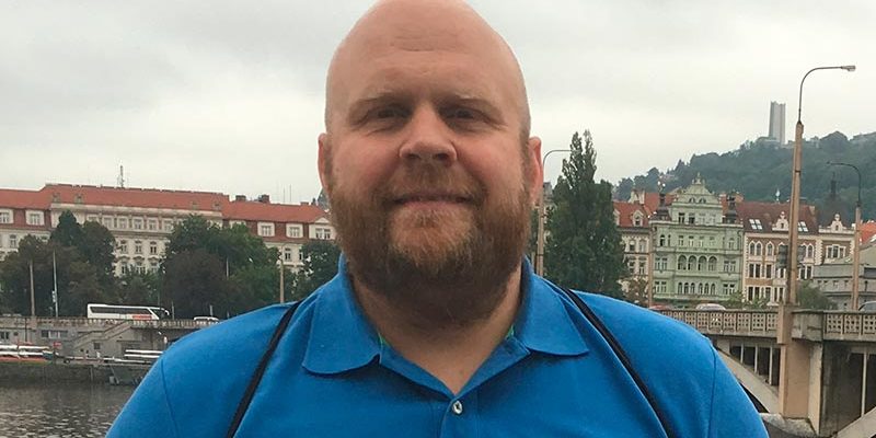 Mies seisoo joen rannalla ja katsoo kameraan. Taustalla näkyy kaupunkia.