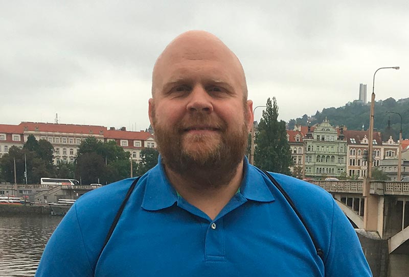 Mies seisoo joen rannalla ja katsoo kameraan. Taustalla näkyy kaupunkia.