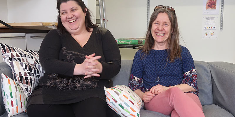 Kaksi naista istuvat sohvalla hymyillen.