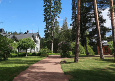 Pihatie, jonka molemmilla puolilla nurmikkoa. Oikealla näkyy vanha tiilirakennus ja vasemmalla vaalea vanha puutalo.