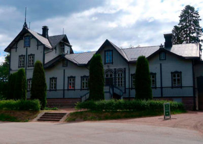 Vanha ja iso vaalea puurakennus. Talon edessä pensasaita ja sypressejä.