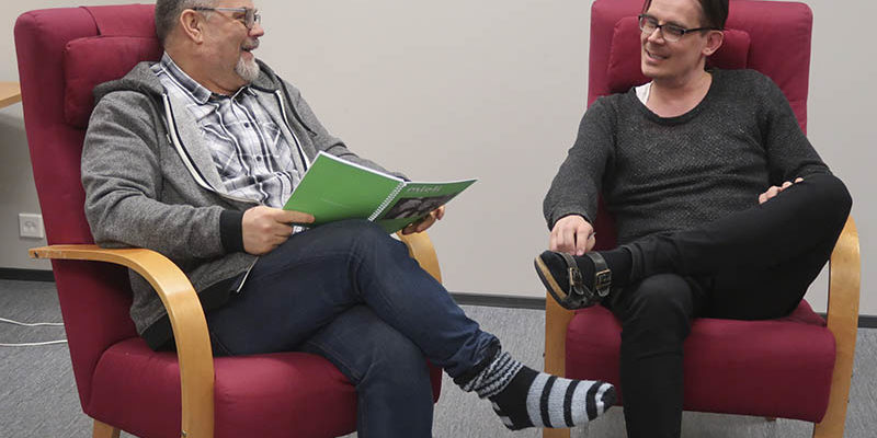 Kaksi miestä istumassa nojatuoleilla. Toinen pitelee kädessään kirjaa.