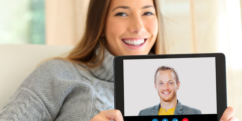Nainen pitelee hymyillen käsissään tablet-laitetta, jonka kuvaruudussa hymyilee mies.