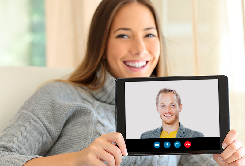 Nainen pitelee hymyillen käsissään tablet-laitetta, jonka kuvaruudussa hymyilee mies.