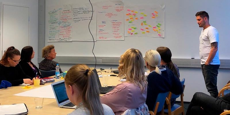 Ihmiset istuvat kokoushuoneessa ja seuraavat opetustilannetta.