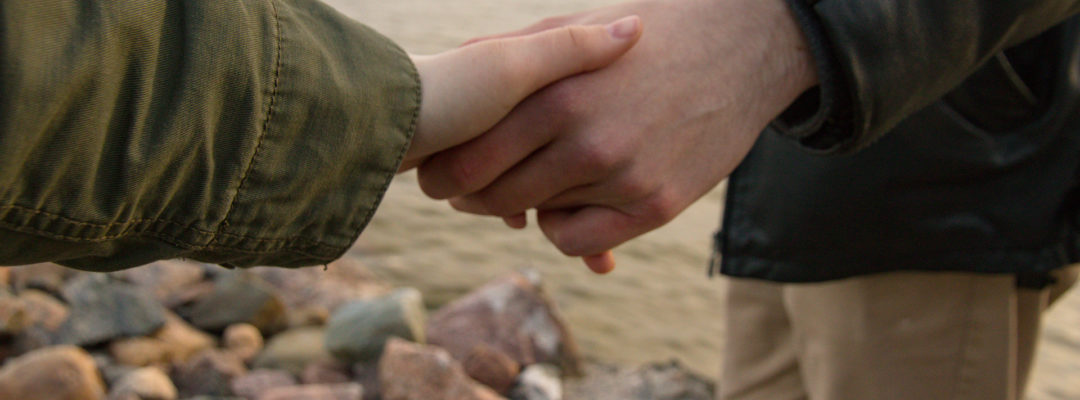 Kaksi henkilöä seisovat rannalla ja pitelevät toisiaan kädestä.