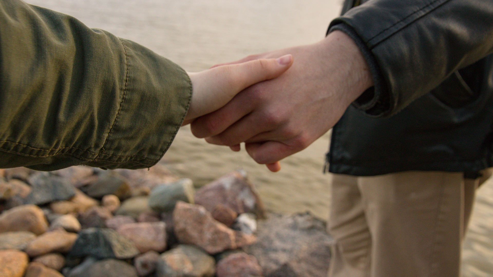 Kaksi henkilöä seisovat rannalla ja pitelevät toisiaan kädestä.