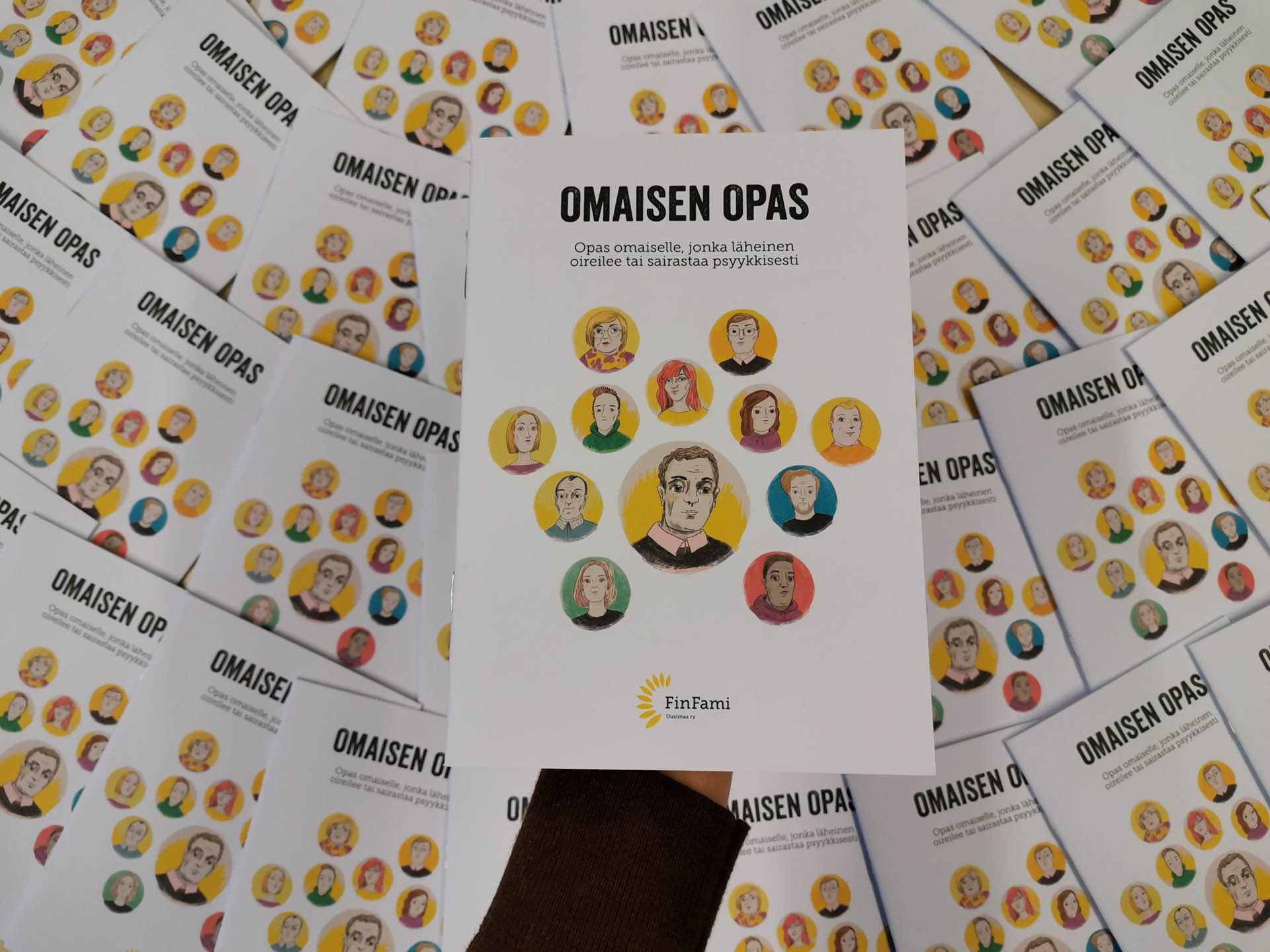 Pöydällä on esillä useita Omaisen opas -vihkoja. Teoksen kannessa lukee "Omaisen opas, opas omaiselle, jonka läheinen oireilee tai sairastaa psyykkisesti."