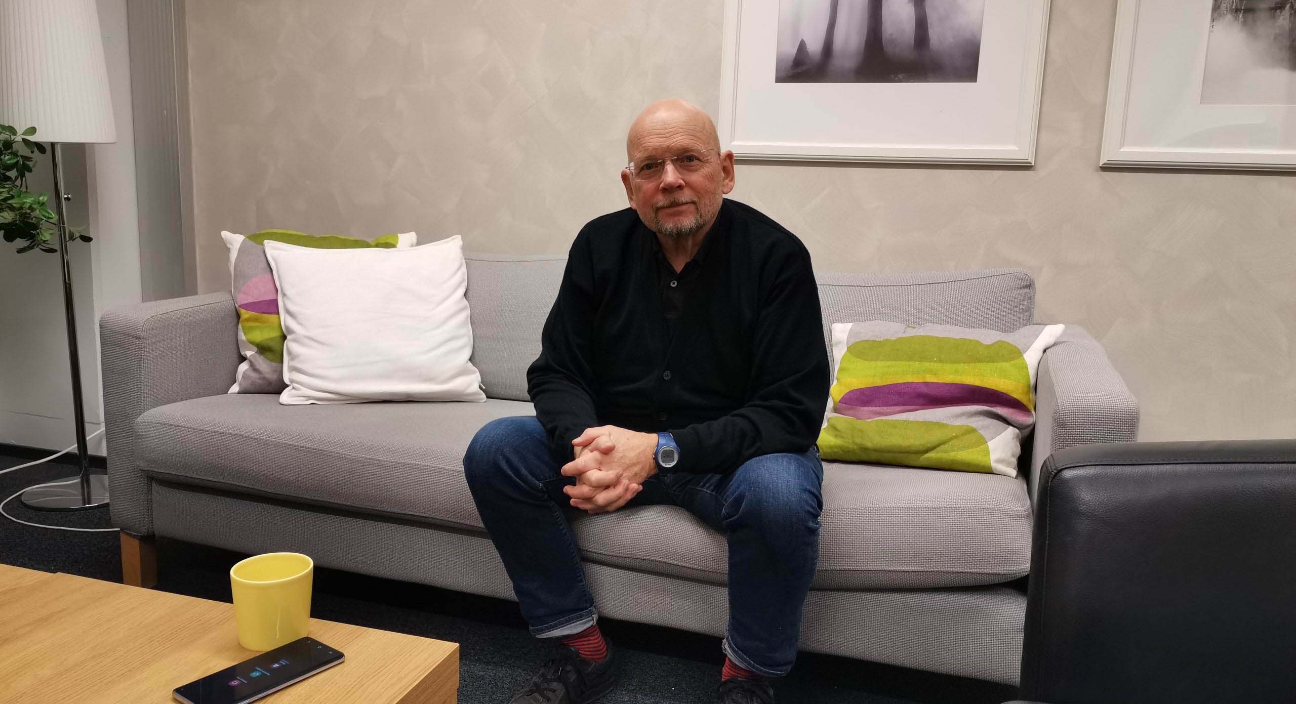 Mies istuu sohvalla kädet ristissä ja katsoo kameraan.