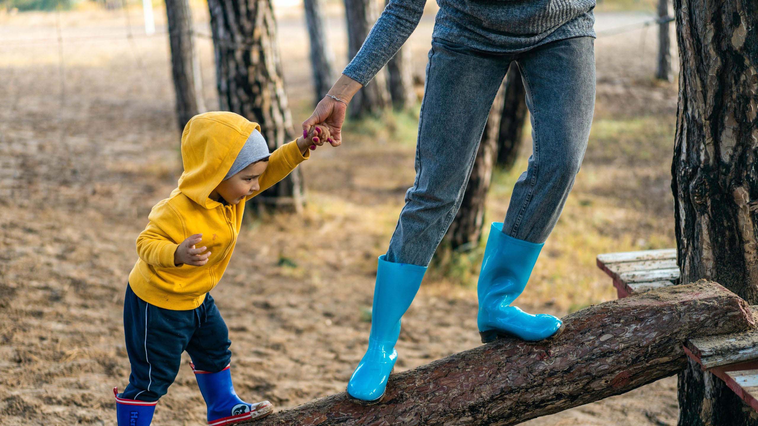 Vanhempi pitää lasta kädestä ja kävelee lapsen kanssa tukin päällä ulkona.