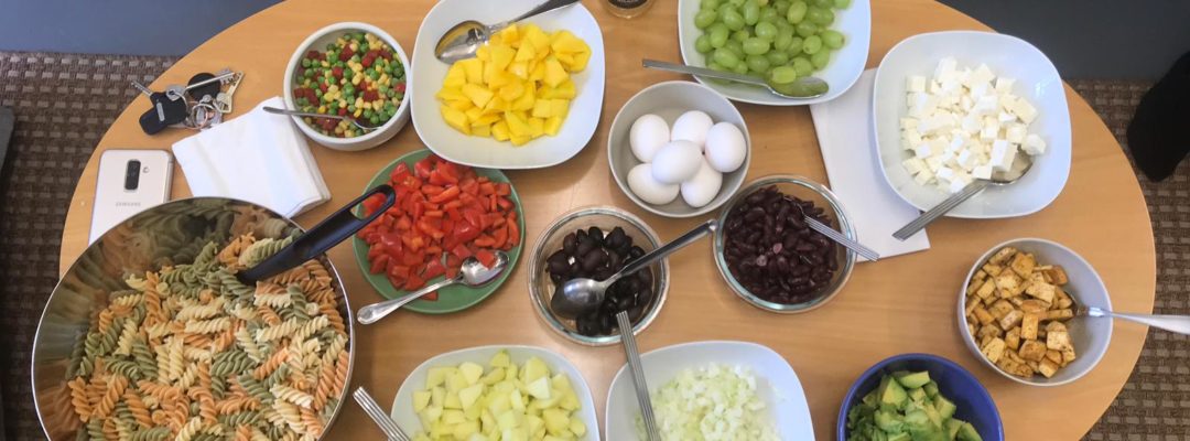 Pöydällä kulhoissa viinirypäleitä, fetajuustoa, oliiveja, kananmunia, ananasta, paprikaa, avokadoa ja pastaa.