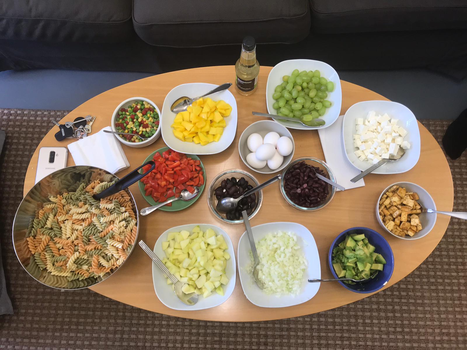 Pöydällä kulhoissa viinirypäleitä, fetajuustoa, oliiveja, kananmunia, ananasta, paprikaa, avokadoa ja pastaa.