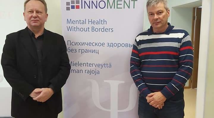 Kaksi miestä seisoo mainostelineen edessä. Mainoksessa lukee: Innoment. Mental health without borders. Psihicheskoe zdorov bez granits. Mielenterveyttä ilman rajoja. www.innoment.pro.