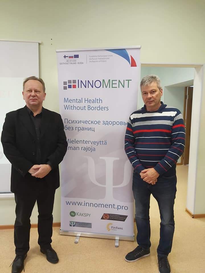 Kaksi miestä seisoo mainostelineen edessä. Mainoksessa lukee: Innoment. Mental health without borders. Psihicheskoe zdorov bez granits. Mielenterveyttä ilman rajoja. www.innoment.pro.