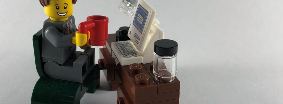 Lego-hahmo istuu tuolilla lego-tietokoneen äärellä punainen kahvikuppi kädessä ja hymyilee.