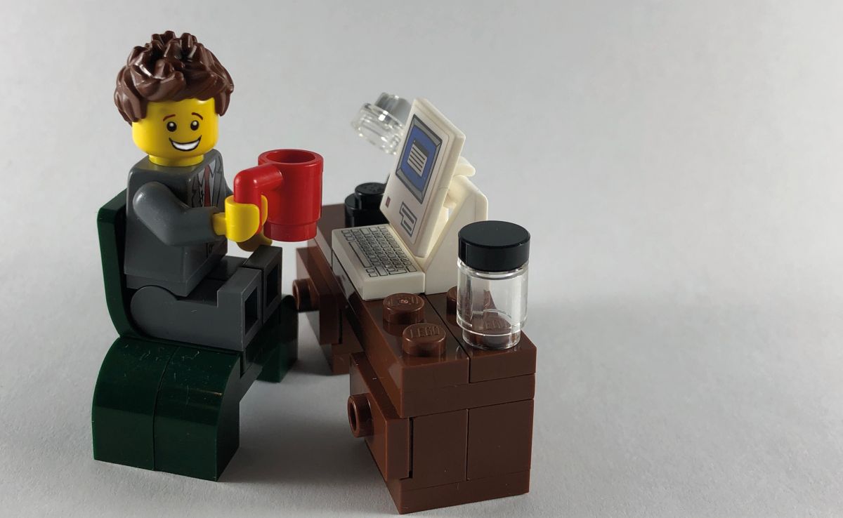 Lego-hahmo istuu tuolilla lego-tietokoneen äärellä punainen kahvikuppi kädessä ja hymyilee.