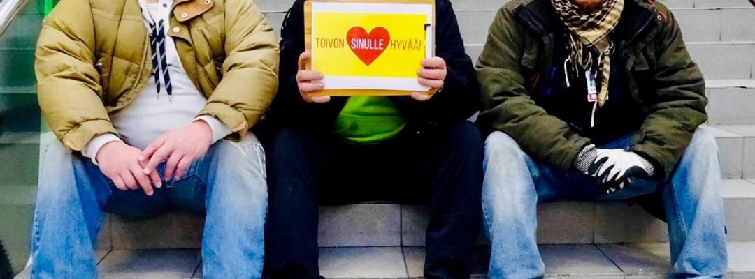 Kolme miestä istuvat portailla. Keskimmäisen käsissä on kyltti, jossa lukee "toivon sinulle hyvää!".