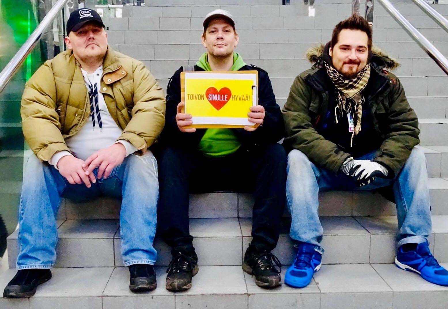 Kolme miestä istuvat portailla. Keskimmäisen käsissä on kyltti, jossa lukee "toivon sinulle hyvää!".