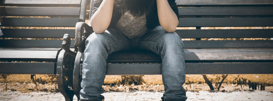 Mies istuu puistonpenkillä pidellen päätään ja katsoo maahan.