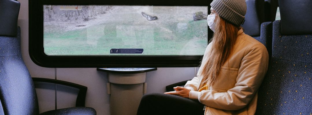 Nainen istuu junassa kasvomaski kasvoilla.