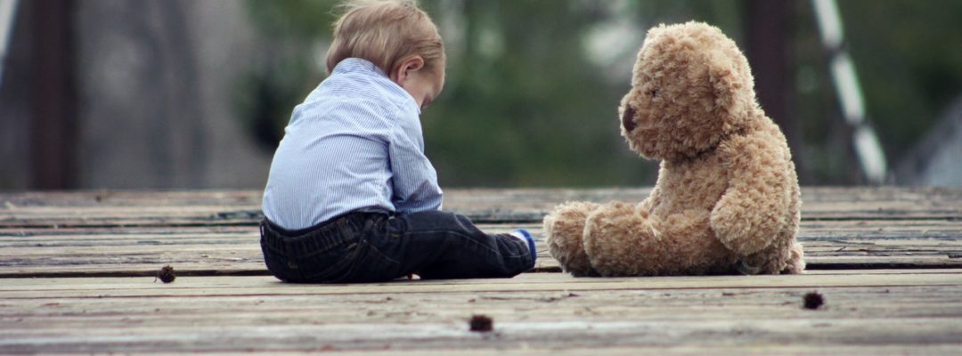 Pieni lapsi istuu maassa nalle vierellään.